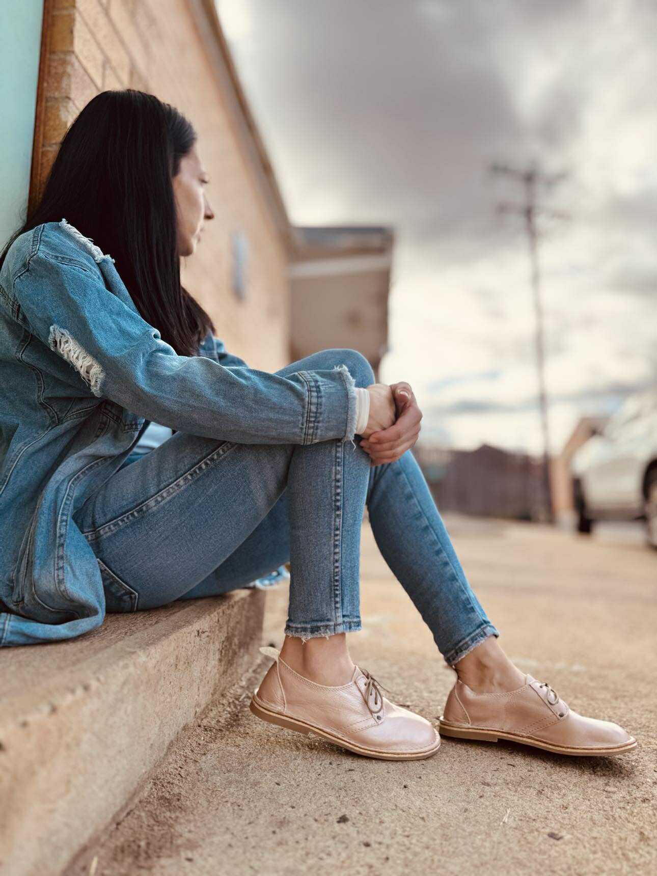 Person sitting on a ledge wearing blue jeans and beige shoes, with a blurred background.