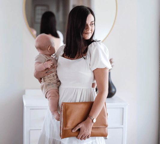 Woman in a white dress holding a baby and a brown Humba Hides Travel Clutch Portable Nappy Changing Station in a room with a mirror.